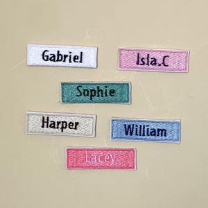 May include: Six rectangular embroidered name patches featuring the names Gabriel, Isla.C, Sophie, Harper, William, and Lacey. Each patch has a distinct background color including white, pink, teal, beige, light blue, and dark pink with dark text.