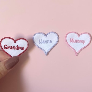 Może przedstawiać: Trzy naszywki z tkaniny w kształcie serca z białymi środkami i haftowanymi brzegami. Pierwsza naszywka ma czerwony brzeg i napis Grandma. Druga ma jasnoniebieski brzeg i napis Nanna. Trzecia ma różowy brzeg i napis Mummy.