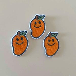 May include: Three embroidered iron-on patches, each shaped like an orange mango with a green leaf at the top. Each mango features a black stitched smiley face with two circular eyes and a curved mouth, all outlined in white thread.