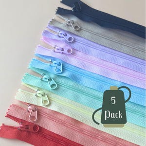 May include: A pack of 5 colorful zipper closures in a rainbow of colors, including red, pink, yellow, green, blue, purple, and gray. The zippers are arranged in a stack, with the red zipper at the bottom and the gray zipper at the top. The text "5 Pack" is visible in the image.