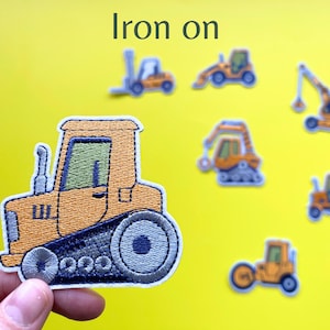 May include: Embroidered iron-on patches of construction vehicles in orange, blue, and yellow. The patches include a tractor, excavator, and other machinery. The text "Iron on" is displayed at the top.