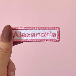 May include: A rectangular embroidered patch featuring a white background with the name Alexandria stitched in pink thread. The patch is bordered by a thick pink stitched edge, creating a clear contrast between the lettering and the frame.