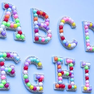 May include: Close-up of alphabet letters A, B, C, D, F, G, H, and I. Each letter is outlined with small rhinestones and filled with colorful beads. The letters are on a light blue background. Ideal for crafts and personalization.