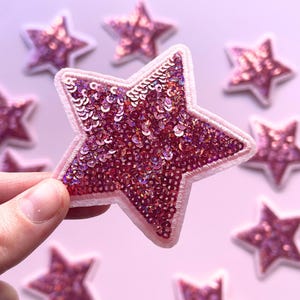 May include: A collection of pink sparkly star-shaped iron-on patches. The patches are made of sequins and have a white outline.