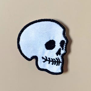 May include: An embroidered patch featuring a white skull with black stitching details. The design includes a black outline, black eye sockets, a small black nose cavity, and a stitched mouth line that resembles a fish bone structure.