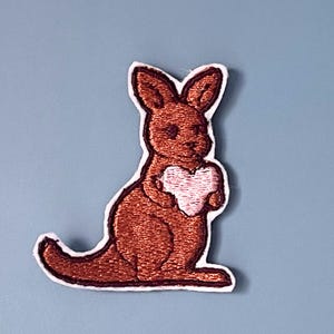 May include: An embroidered patch featuring a brown kangaroo sitting in profile. The kangaroo has dark brown stitched details and is holding a light pink heart shape against its chest. The entire design is outlined with a thick white border.