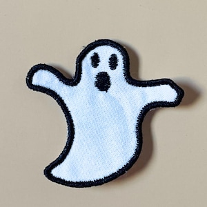 May include: A white ghost-shaped embroidered patch with a thick black stitched border. The ghost features two oval black eyes and a round black mouth, creating a simple, high-contrast design with a textured fabric surface and defined edges.