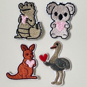 May include: Four embroidered patches of Australian animals. An alligator, koala, and kangaroo each hold a pink heart. An ostrich holds a red heart. All patches have a white border.