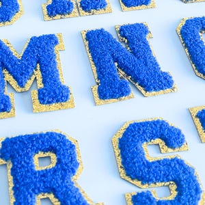 May include: Close-up of several blue chenille letter patches with gold borders. The letters are in a bold, block style, and the blue chenille has a textured, fuzzy appearance. The patches are arranged on a light blue surface.