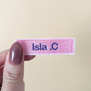 May include: A rectangular, pink embroidered patch with a white border. The text 'Isla .C' is stitched in the centre in a dark purple, sans-serif font. The patch has a textured fabric appearance with visible thread details throughout the design.
