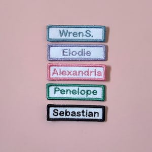May include: Five rectangular embroidered name patches arranged vertically. The patches feature white backgrounds with coloured borders and matching thread for the names: Wren S. in blue, Elodie in lavender, Alexandria in pink, Penelope in green, and Sebastian in black.