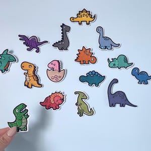 May include: A collection of colorful dinosaur patches in various shapes and sizes. The patches feature embroidered details and a variety of colors, including green, orange, blue, and purple. The patches are designed for decorative purposes.