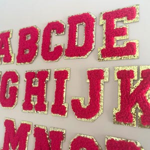 May include: Close-up of a collection of red, fuzzy, varsity-style letter patches with gold glitter borders. The letters are arranged in rows, showcasing the alphabet. Ideal for personalized crafts and apparel.