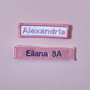 May include: Two rectangular embroidered name patches. The top patch has a white background with pink border and the name Alexandria in pink thread. The bottom patch is solid pink with a white border and the name Ellana 3A in dark purple thread.