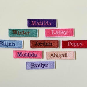 Peut inclure: Plusieurs écussons brodés avec des noms, dans des couleurs variées comme le bleu, le rose, le rouge et le marron. Chaque écusson présente un nom différent brodé avec un fil de couleur contrastante. Noms : Matilda, Elijah, Winter, Lacey, Jordan, Poppy, Abigail, et Evelyn.