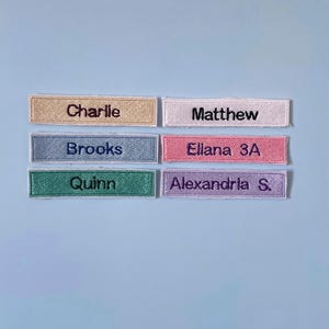May include: Six rectangular embroidered name patches in various colours. The patches feature the names Charlie, Matthew, Brooks, Ellana 3A, Quinn, and Alexandria S. stitched in dark thread on light-coloured fabric backgrounds with finished edges.