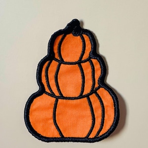 May include: An embroidered patch featuring a stack of three orange pumpkins. The design is outlined in thick black thread, with black stitching defining the segments of each pumpkin. The patch has a flat, fabric-like texture and a distinct shape.