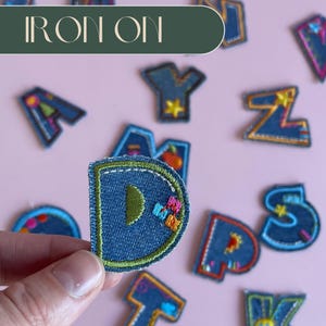 Może przedstawiać: Zestaw naszywek z alfabetem do prasowania wykonanych z dżinsu z kolorowymi haftami. Naszywki są w różnych literach, w tym D, Y, Z, W i S. Naszywki znajdują się na różowym tle z napisem "IRON ON" w zielonym prostokącie u góry obrazu.