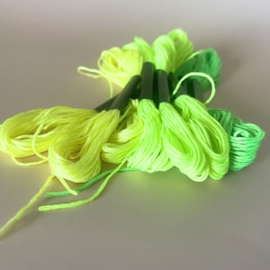 May include: A selection of neon green and yellow embroidery floss skeins. The floss is arranged in a fan shape, with the colours transitioning from yellow to bright green. The skeins are tightly wound and ready for use in various craft projects.
