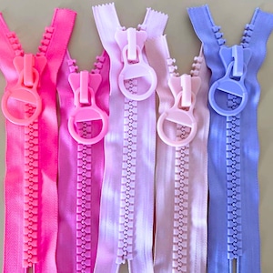 May include: Close-up of five colorful zippers in shades of pink and purple. Each zipper has a matching pull tab and teeth. The zippers are arranged vertically, showcasing their open design and vibrant hues, perfect for sewing projects.