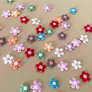 以下が含まれることがあります： ピンク、オレンジ、黄色、赤、青、緑、茶色など、さまざまな色の小さな刺繍の花のアップリケのコレクション。 花は薄茶色の表面に散らばっており、装飾的なディスプレイを作成しています。