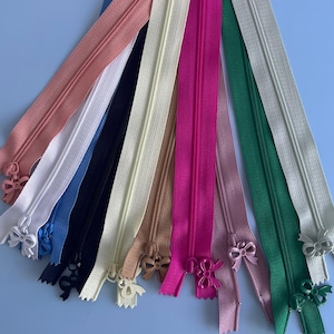 Może przedstawiać: Kolekcja kolorowych zamków błyskawicznych w różnych odcieniach, w tym różowym, białym, niebieskim, czarnym, beżowym, brązowym, fuksji i zielonym. Każdy zamek ma dekoracyjny uchwyt w kształcie kokardy. Zamki są ułożone na jasnoniebieskim tle.