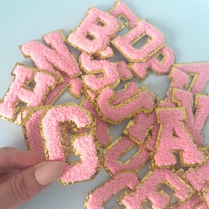May include: A selection of pink, fluffy, embroidered letters with gold edging. The letters are in different fonts and sizes, suitable for crafting or customisation. The background is a pale blue.