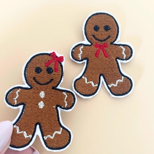 May include: Two embroidered gingerbread man patches. One has a red bow, the other a red bow tie. Both have black eyes and smiles, with white zig-zag trim and a white border. The patches are brown and are on a light beige background.