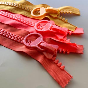 以下が含まれることがあります： 黄色、コーラル、オレンジの色合いの、3つのカラフルなジッパーのクローズアップ。各ジッパーには、大きくて丸いプルタブが付いています。ジッパーの歯が見え、生地は質感があります。