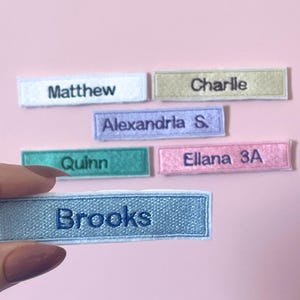 May include: A collection of rectangular embroidered name patches in various colours including white, gold, light purple, teal, pink, and light blue. Each patch features a name stitched in a dark thread, specifically Matthew, Charlie, Alexandria S., Quinn, Ellana 3A, and Brooks.