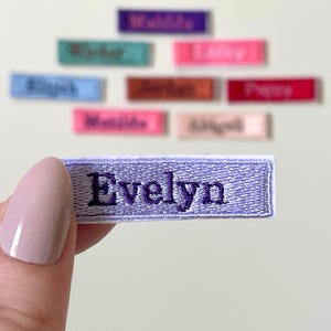 May include: A lavender embroidered name patch with the name "Evelyn" in dark purple thread. Other name patches in various colours are in the background.