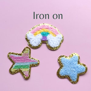 May include: Three iron-on patches: a rainbow with clouds, a pink and green striped star, and a blue star. The patches are on a pink background. Iron on.