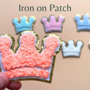 May include: Iron-on patch set of 6 fuzzy crown shapes in pink, blue, white, and purple. The crowns are outlined in gold.