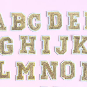 Peut inclure: Lettres de l'alphabet A à P en sequins dorés, bordées de blanc, disposées sur un fond rose clair. Les lettres sont de style bloc classique, idéales pour les projets de personnalisation.