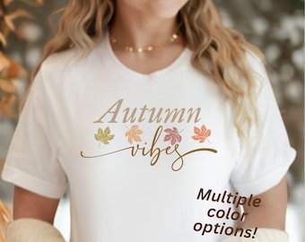May include: White t-shirt with the words "Autumn vibes" in a brown script font, decorated with colorful leaf graphics. The text "Multiple color options!" is also visible. The shirt is a casual, seasonal apparel item.