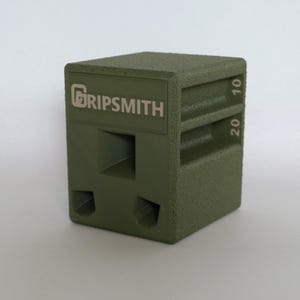 May include: Olive green cube-shaped object with the word "GRIPSMITH" printed on the side. The cube has textured surfaces and cut-out shapes. Numbers 10 and 20 are printed on the side. The object appears to be made of a solid material.