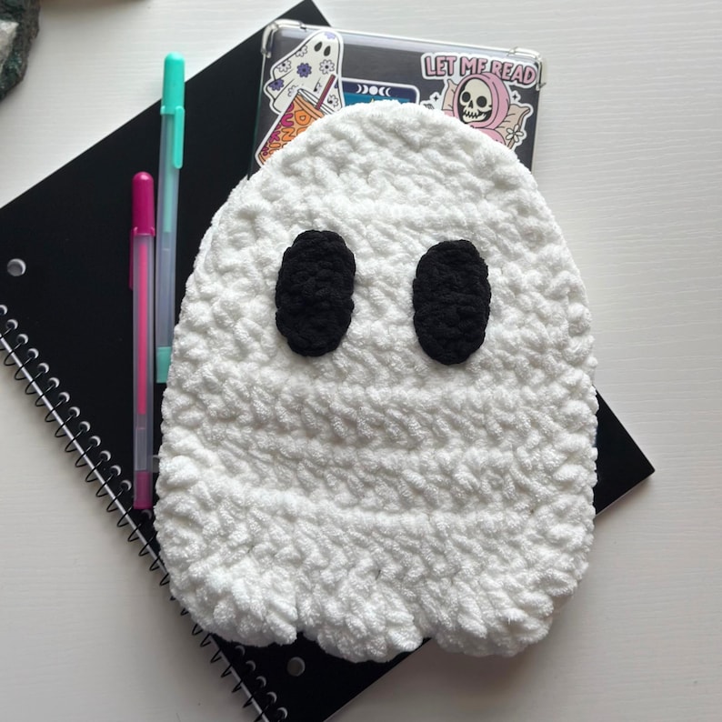 May include: A white, crocheted ghost with black oval eyes, resting on a black spiral notebook. Two pens are placed beside the ghost. A phone case with a skull sticker that says "LET ME READ" is also visible.
