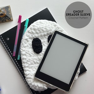 May include: A white crochet ghost e-reader sleeve with black eyes, holding an e-reader. The sleeve is on top of a black spiral notebook with pink and teal pens. The text on the image says "GHOST EREADER SLEEVE Crochet Pattern."