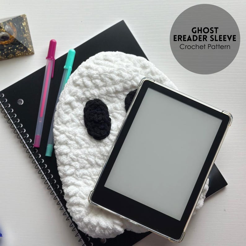 May include: A white crochet ghost e-reader sleeve with black eyes, holding an e-reader. The sleeve is on top of a black spiral notebook with pink and teal pens. The text on the image says "GHOST EREADER SLEEVE Crochet Pattern."