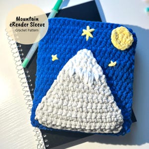 May include: A blue crocheted eReader sleeve featuring a white mountain, yellow stars, and a yellow moon. The sleeve is made with a fluffy yarn and has a crocheted texture. The text "Mountain eReader Sleeve Crochet Pattern" is at the top of the sleeve.