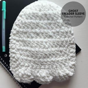 May include: A white, crocheted eReader sleeve with a ghost-like shape. The sleeve is made of a textured, fluffy yarn. A teal pen and a black spiral notebook are visible in the background. The text "GHOST EREADER SLEEVE Crochet Pattern" is on a circular label.