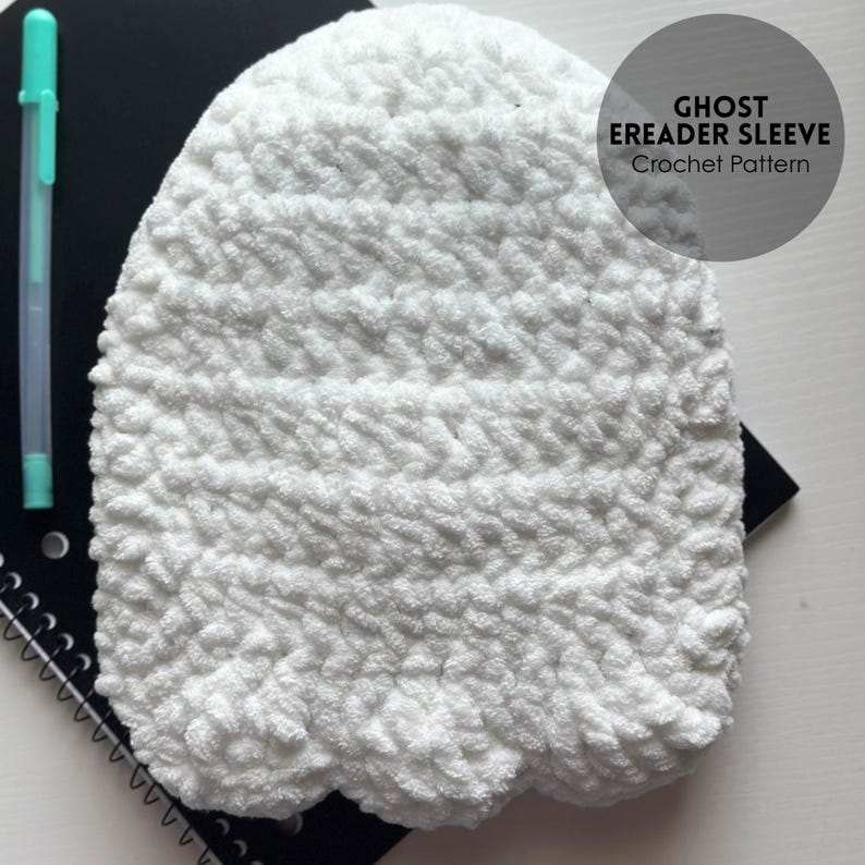 May include: A white, crocheted eReader sleeve with a ghost-like shape. The sleeve is made of a textured, fluffy yarn. A teal pen and a black spiral notebook are visible in the background. The text "GHOST EREADER SLEEVE Crochet Pattern" is on a circular label.