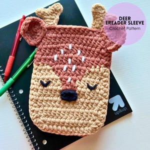 May include: A crocheted eReader sleeve in a deer design, featuring shades of brown, beige, and white. The sleeve has a deer face with antlers, eyes, and a nose. Text in a pink circle reads "DEER EREADER SLEEVE Crochet Pattern".