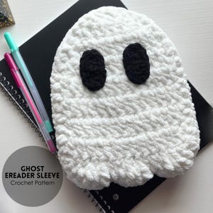 May include: A white, crocheted ghost eReader sleeve with black oval eyes. The sleeve is on a black spiral notebook with pens. The text "GHOST EREADER SLEEVE Crochet Pattern" is in a circle on the bottom left.