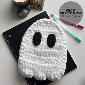 May include: A white, crocheted ghost eReader sleeve with black oval eyes. The sleeve is on a black spiral notebook. The text "GHOST EREADER SLEEVE Crochet Pattern" is in a circle in the upper right corner. Two pens are next to the notebook.
