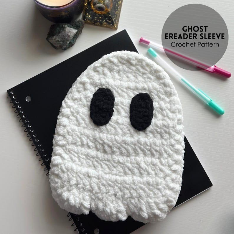 May include: A white, crocheted ghost eReader sleeve with black oval eyes. The sleeve is on a black spiral notebook. The text "GHOST EREADER SLEEVE Crochet Pattern" is in a circle in the upper right corner. Two pens are next to the notebook.