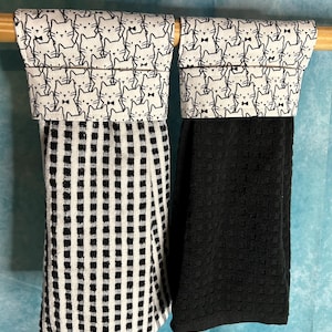May include: Two hanging kitchen towels. One towel is black and white checkered, and the other is solid black. Both towels have a top section with a repeating pattern of black and white cat faces with bow ties.