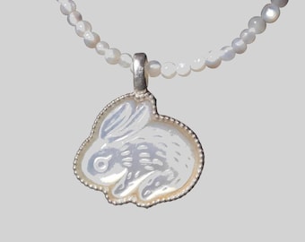 Peut inclure: Un collier avec un pendentif en nacre blanche irisée en forme de lapin. Le pendentif est entouré d'un cadre en métal argenté avec un bord perlé. La chaîne du collier est composée de petites perles rondes en nacre blanche irisée.