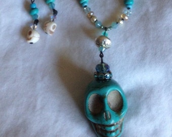 May include: A turquoise skull pendant necklace with a beaded chain. The necklace features a large turquoise skull pendant, small white skull charms, and various blue and silver beads. The necklace is displayed against a white background.