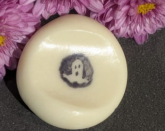 May include: A round, off-white soap with a small, dark gray ghost illustration in the center. The soap is surrounded by purple flowers with yellow centers, creating a Halloween theme.
